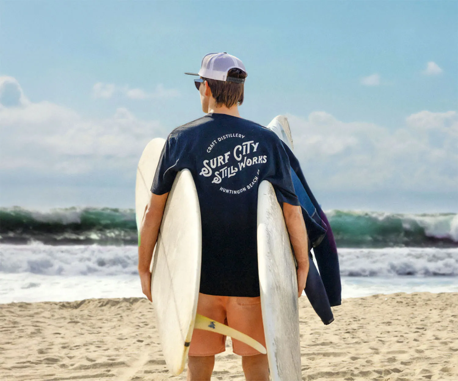Surfer carrying a board on the beach in a Surf City Still Works tee.