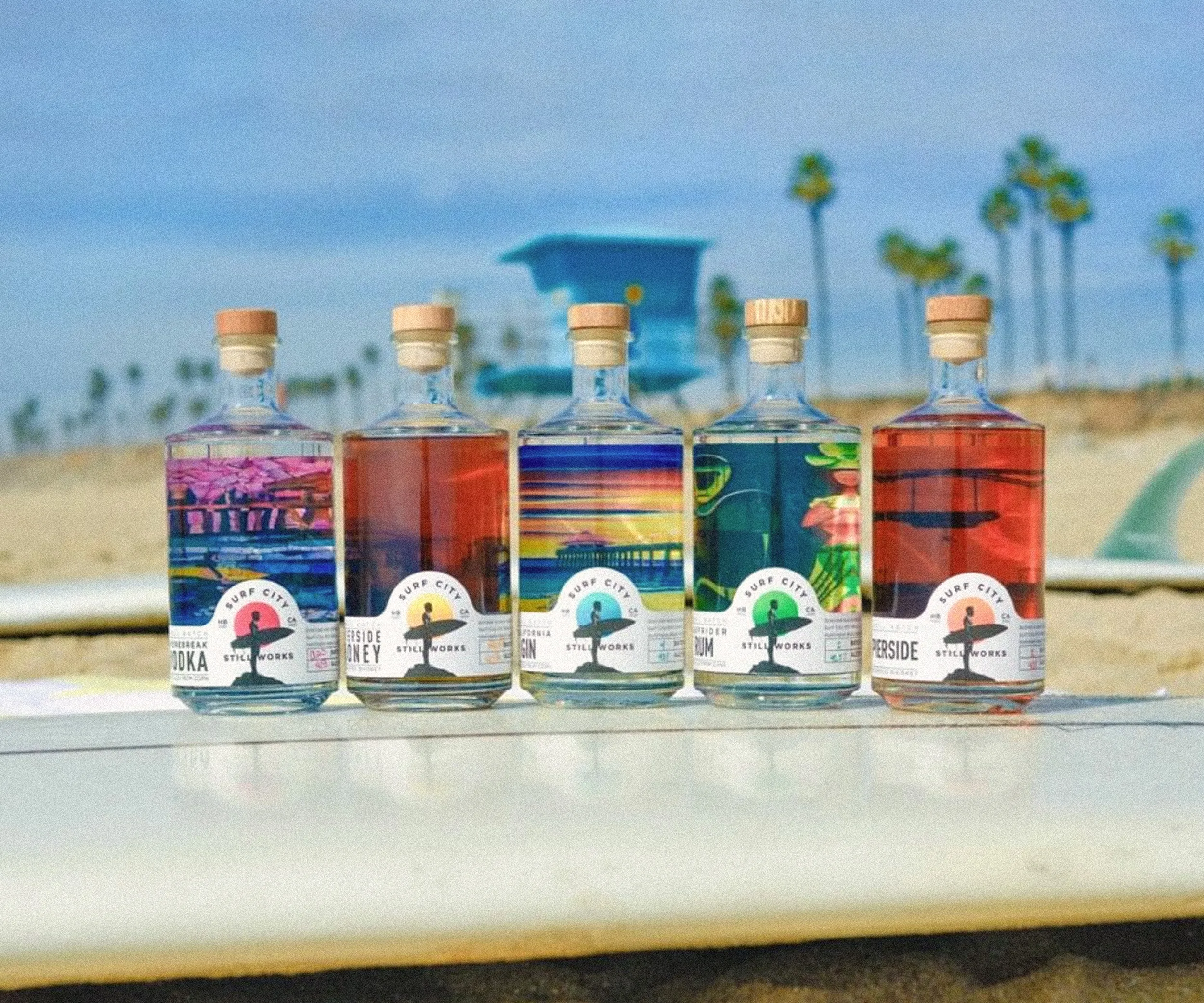 Five spirit bottles lined up on a surfboard with palm trees behind.