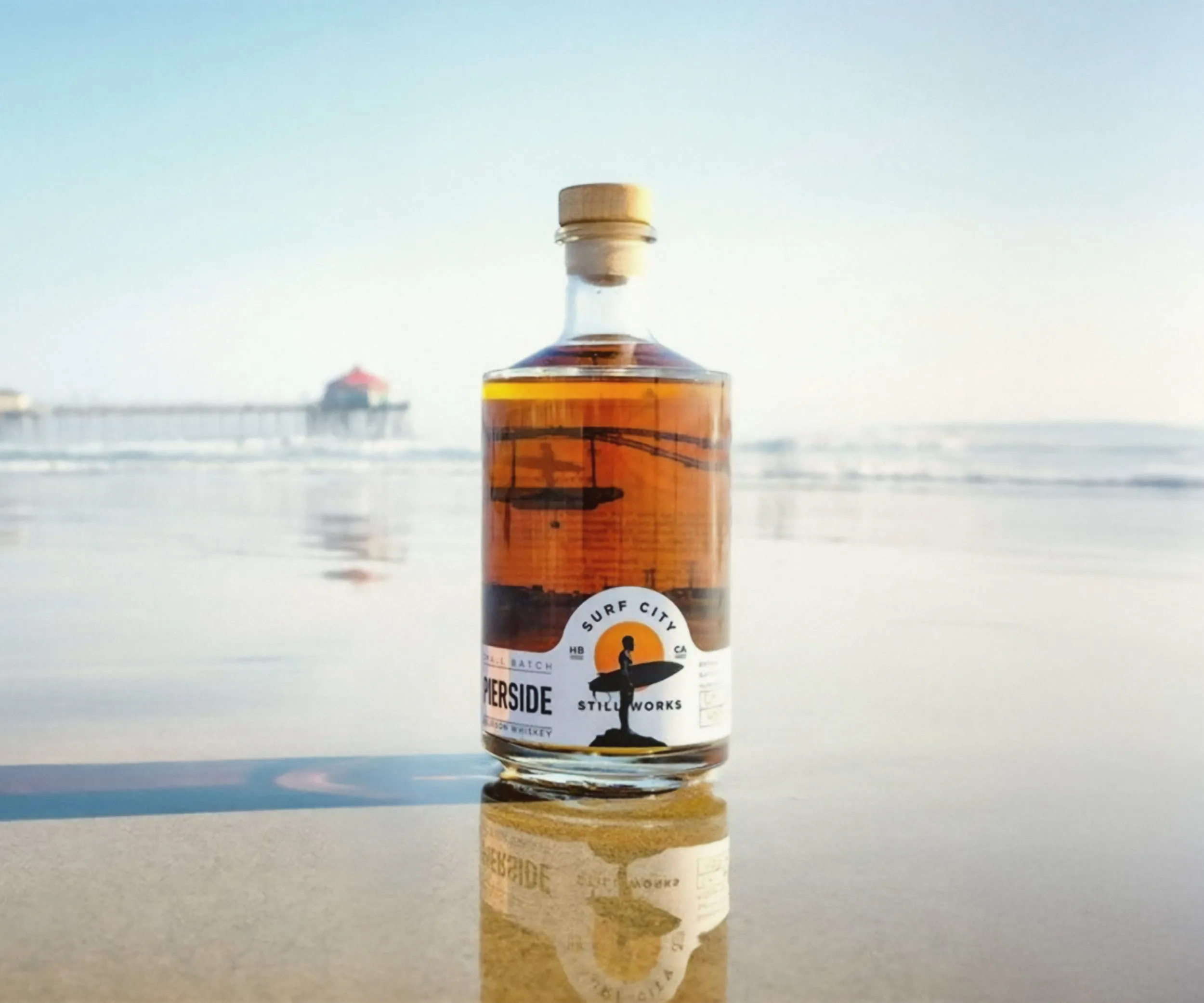 Whiskey bottle on wet sand with the Huntington Beach pier behind.